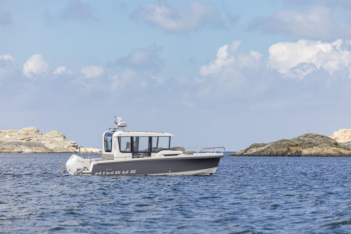 A Nimbus boat navigates calm waters near rocky shores under a blue sky with clouds, showcasing its sleek design.
